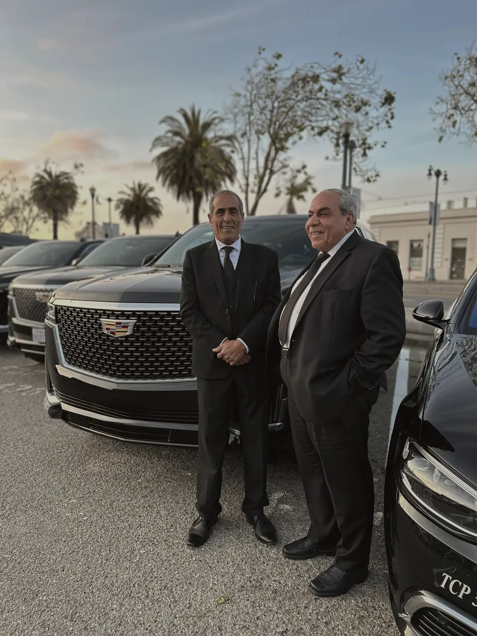 2 BlackWingz chauffeurs in dark suits beside a black Cadillac Escalade