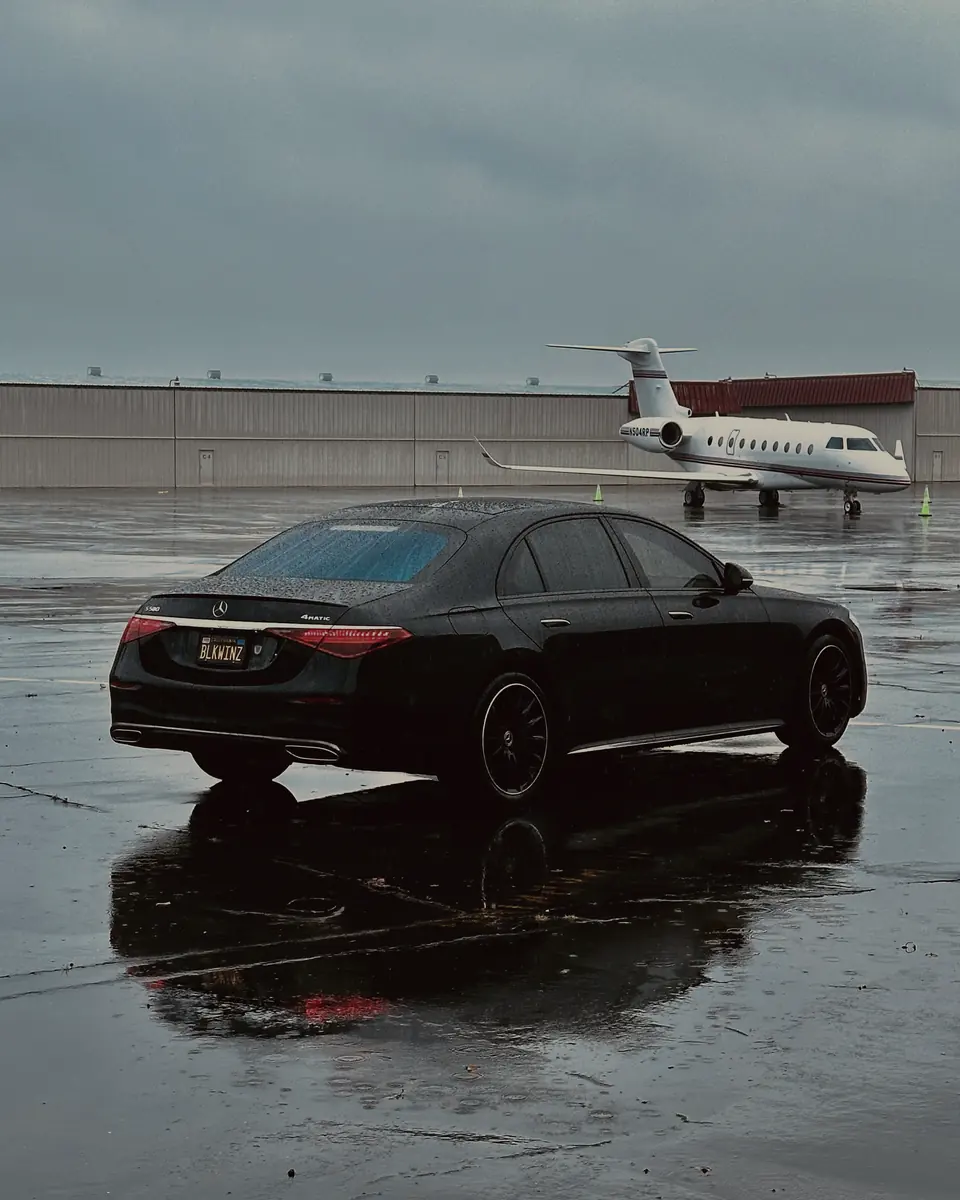 Black Mercedes S-Class parked on a wet ramp, a private jet behind it