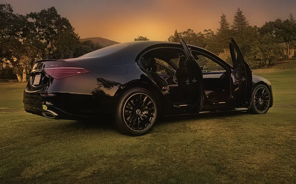 A black Mercedes S-Class with its doors open on grass at dusk, hills behind it