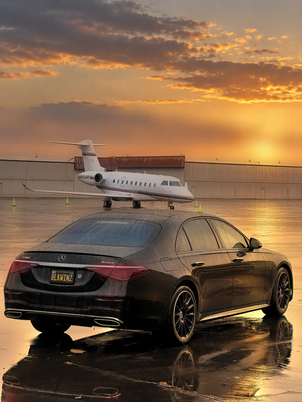 Rear view of a black Mercedes S-Class on a wet ramp at sunset, a private jet beyond it
