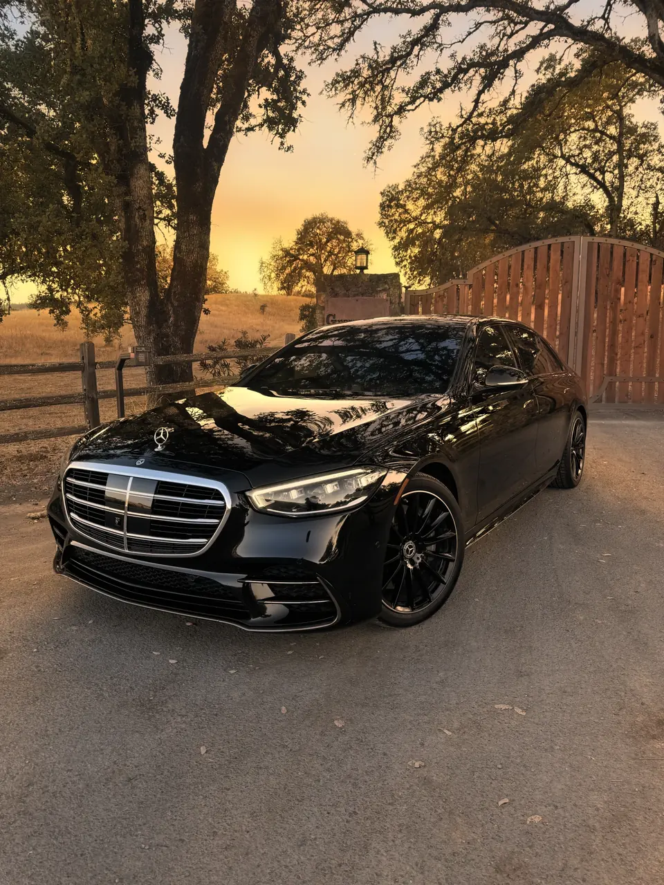 A black Mercedes S-Class beside a wooden ranch gate at golden hour